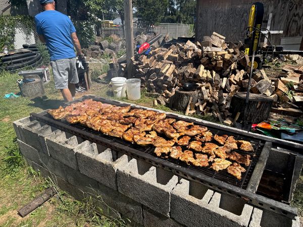 A long outdoor grill full of chicken with firewood stacked behind it